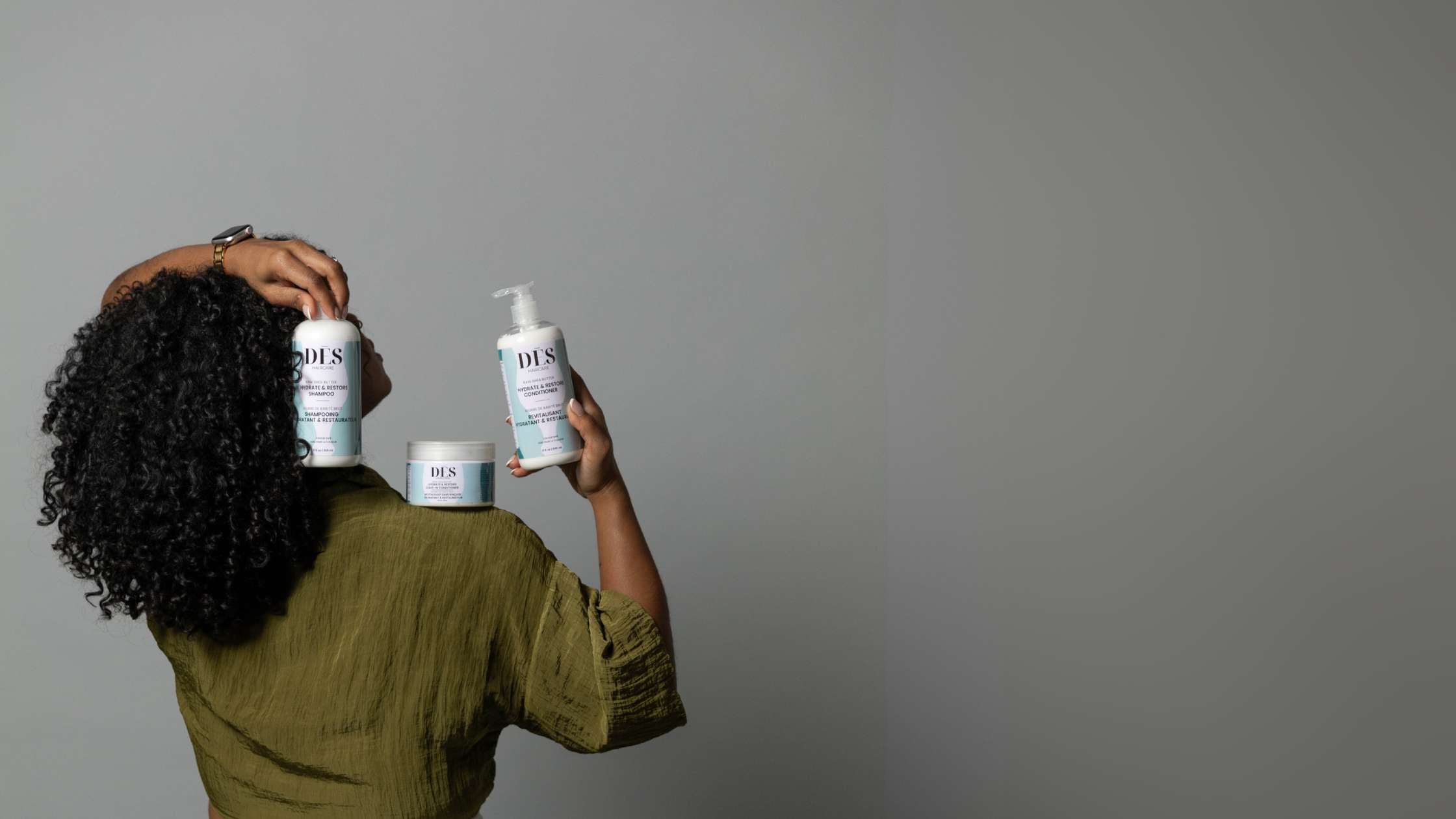 A woman with curly hair holds two bottles of hair care products, including the Volume EdgeLift Brush, smiling confidently.