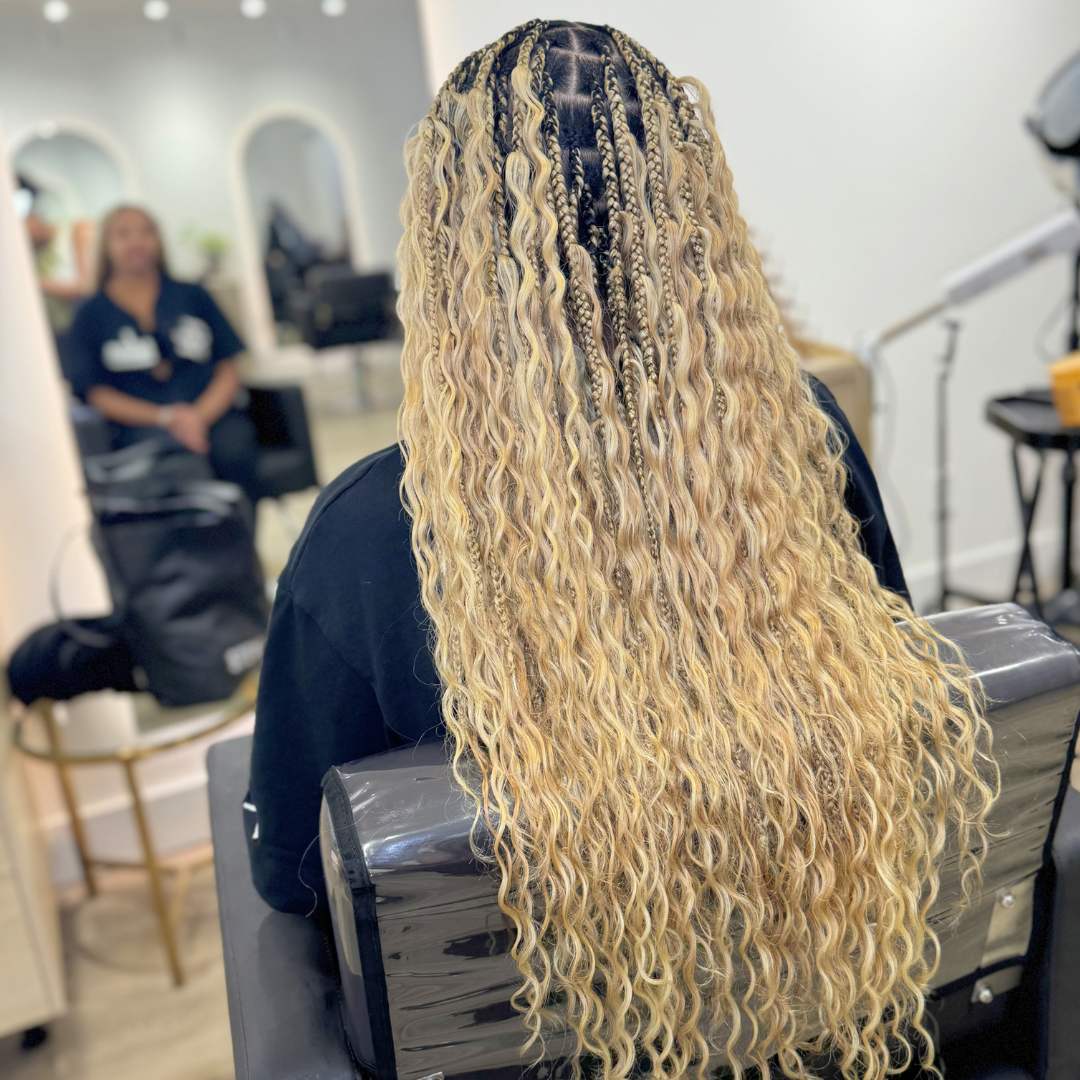 A woman with long, braided curly blonde hair is sitting in a chair, appearing relaxed.