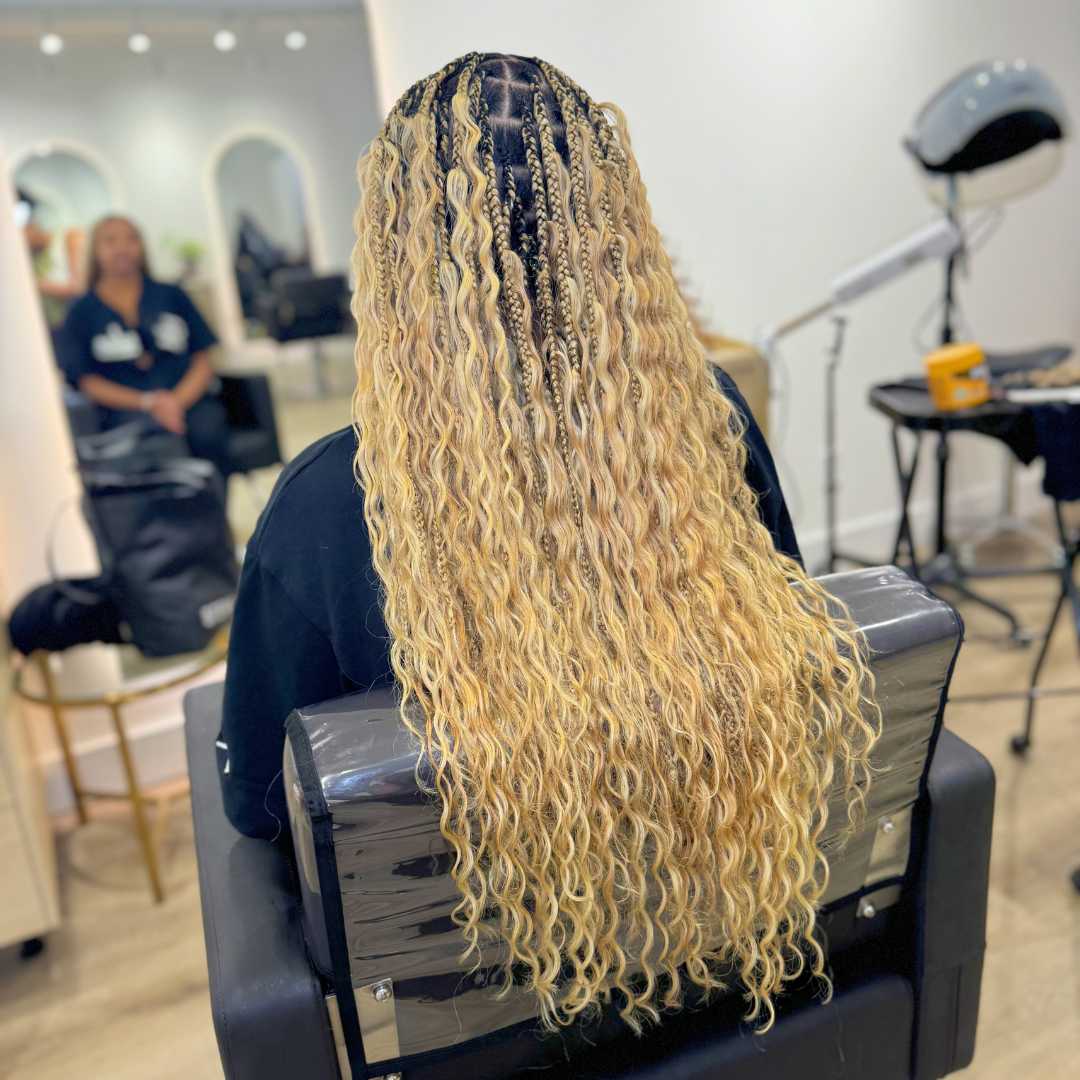 A woman with long, curly blonde hair styled in braids is seated in a chair.