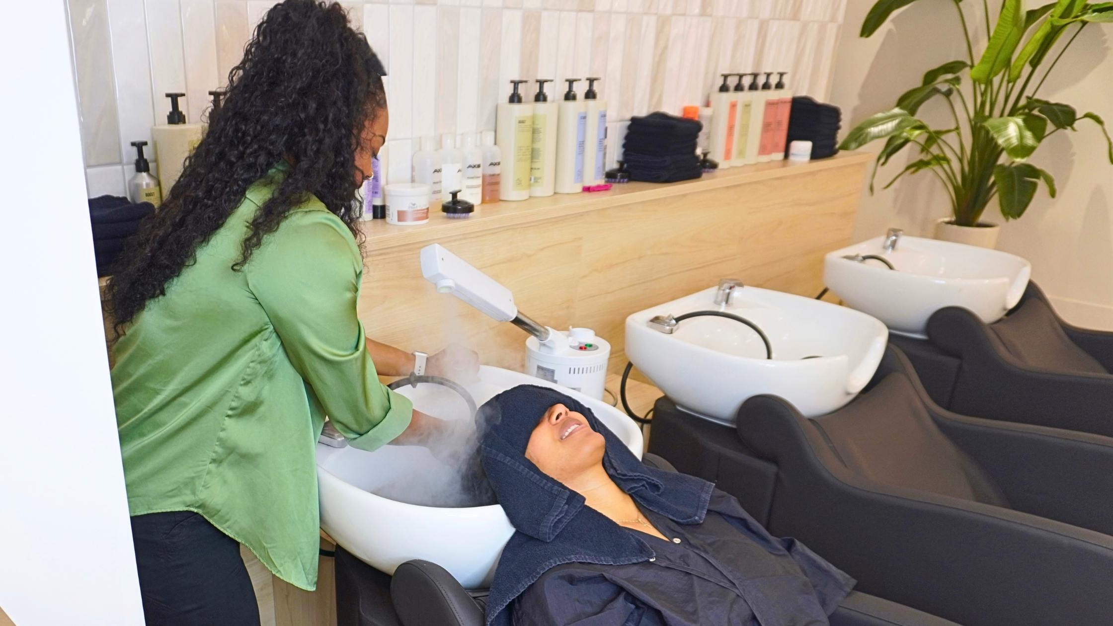 A woman with curly hair braids is having her hair washed at a salon, relaxing in a salon chair.