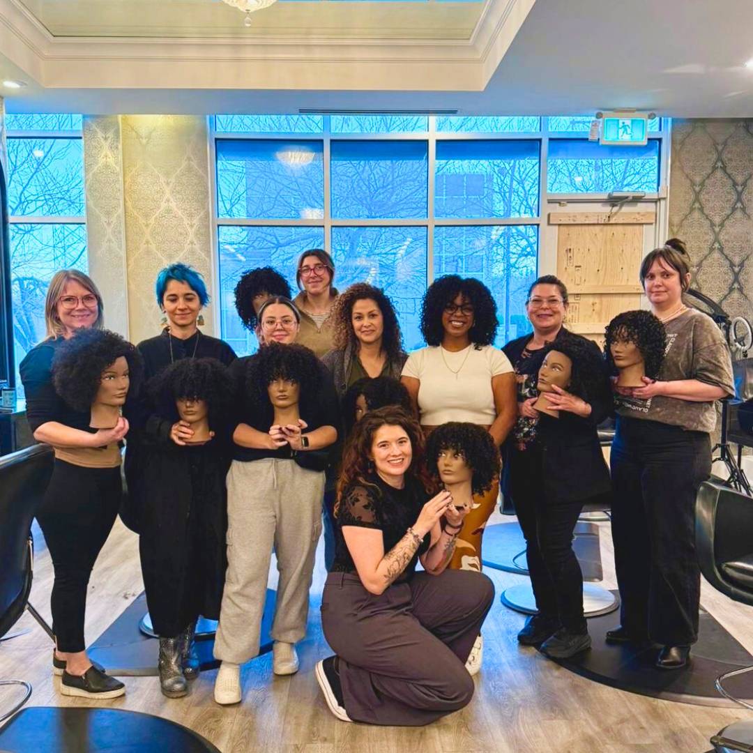 The DES Curl Spa Salon team proudly holding a variety of curly hair wigs, showcasing different textures and styles.