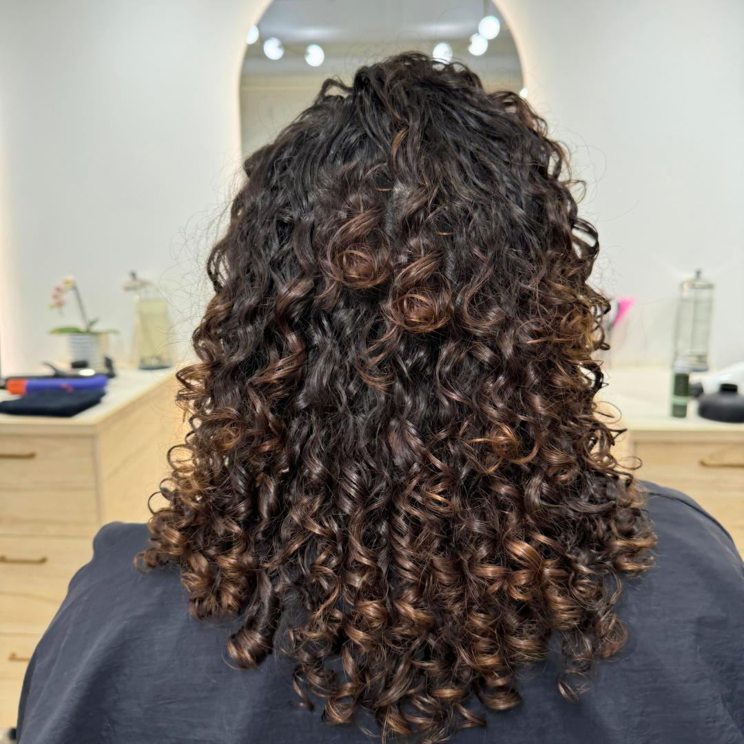 A woman with healthy long curly hair styled in braids sits comfortably in a chair, looking relaxed and content.
